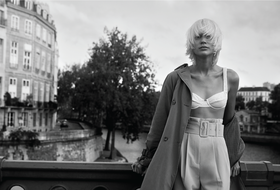 Black-and-white editorial shot: a platinum-blonde model in a trench coat posed on a Parisian bridge with classical architecture in the background.