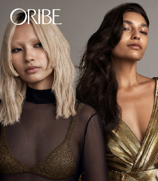 ORIBE brand campaign: two models — a blonde and a brunette — back-to-back in metallic and sheer evening wear with the Oribe wordmark at top left.